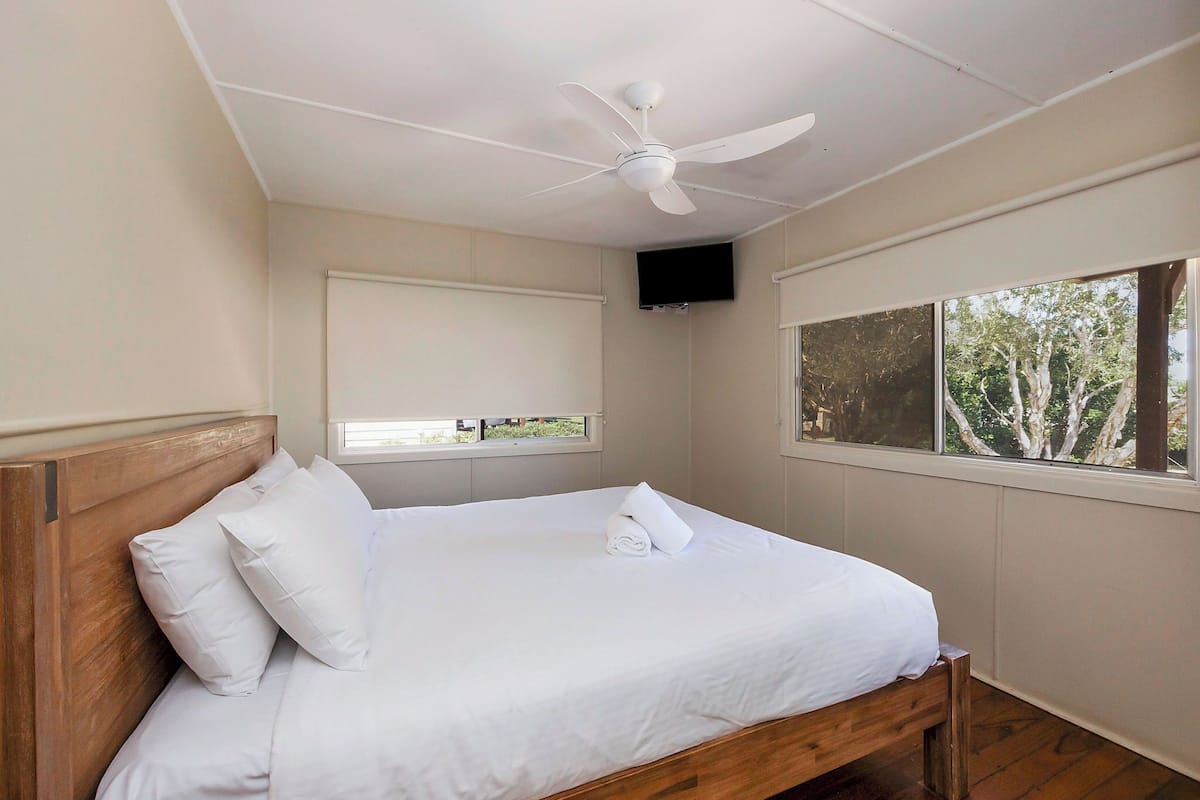 A bedroom with a wooden bed, white bedding, a ceiling fan, a TV mounted on the wall, and a window with a view of trees.