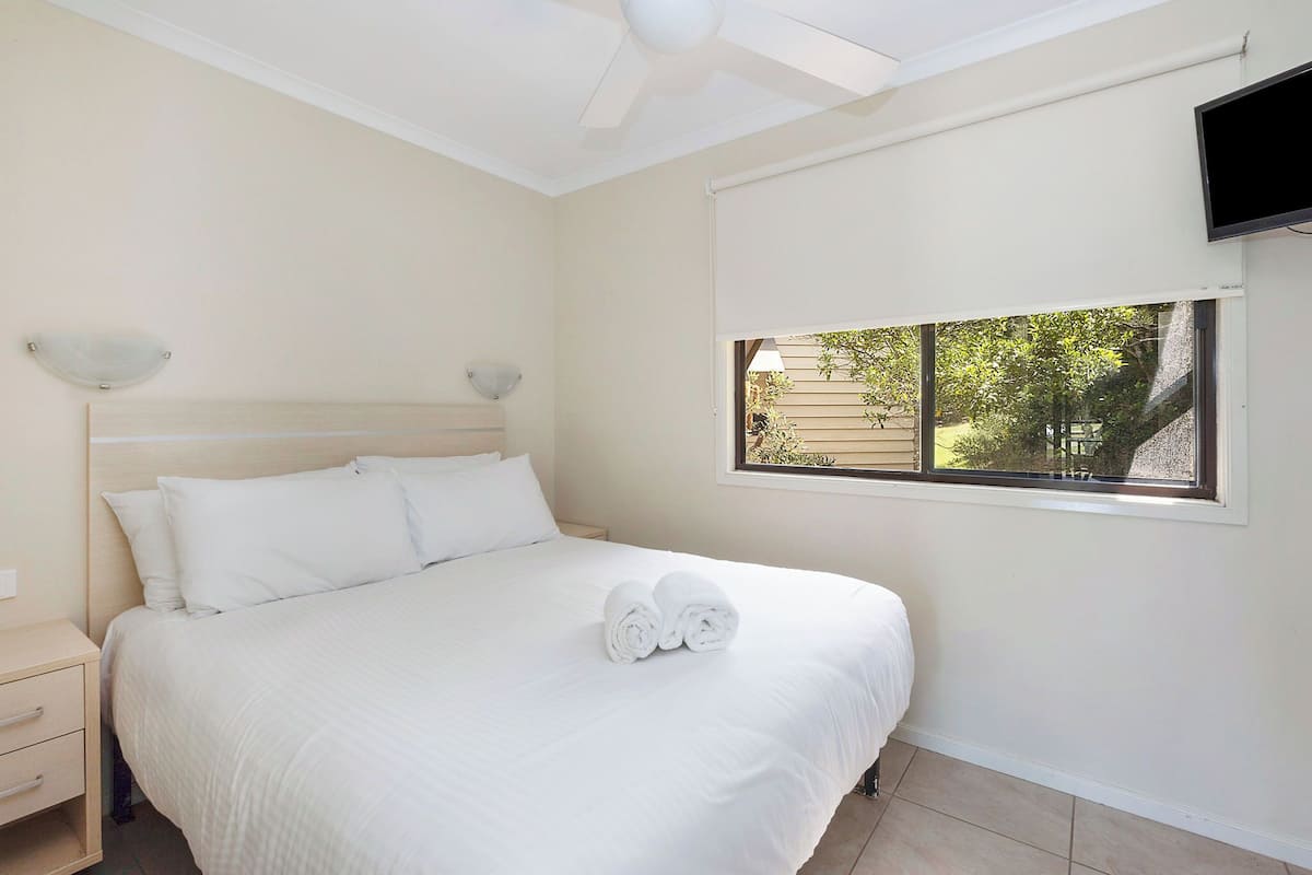 A neatly made bed with white linens and two rolled towels, a bedside table with a lamp, and a window with a view of greenery outside.