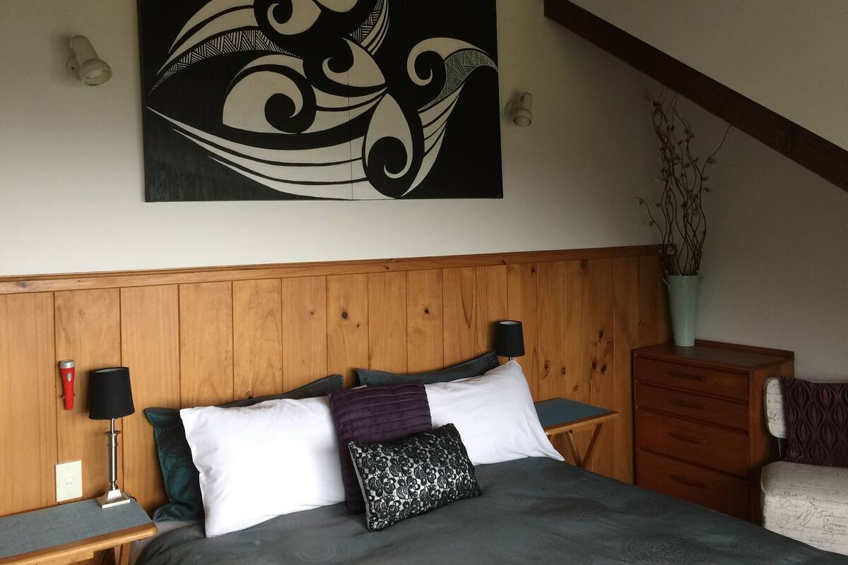 Un dormitorio con cabecera de madera, cama con múltiples almohadas, mesita de noche con lámpara, cómoda y una obra de arte colgada en la pared.