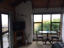 Una sala de estar con televisor sobre un soporte de madera, una mesa de comedor con sillas y ventanas amplias que ofrecen vistas al follaje.