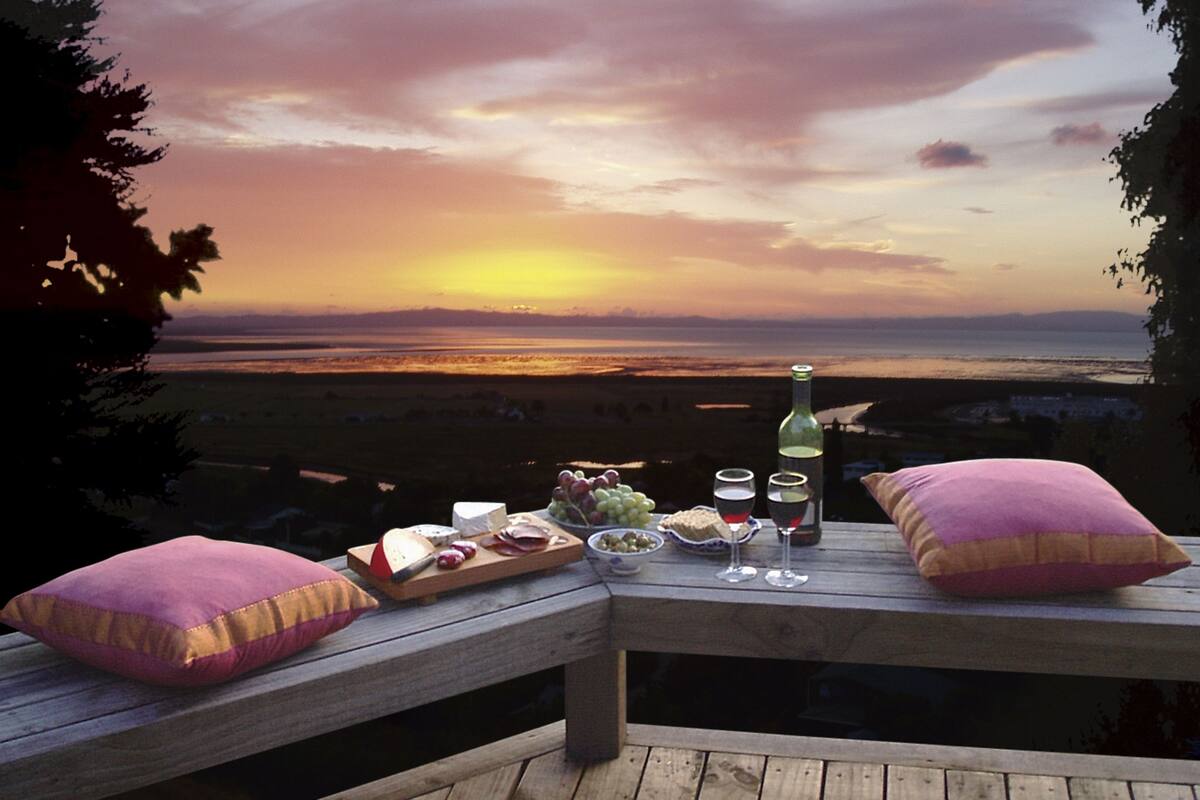 Una terraza de madera con una mesa dispuesta para una comida, con vista a la puesta de sol sobre el agua.