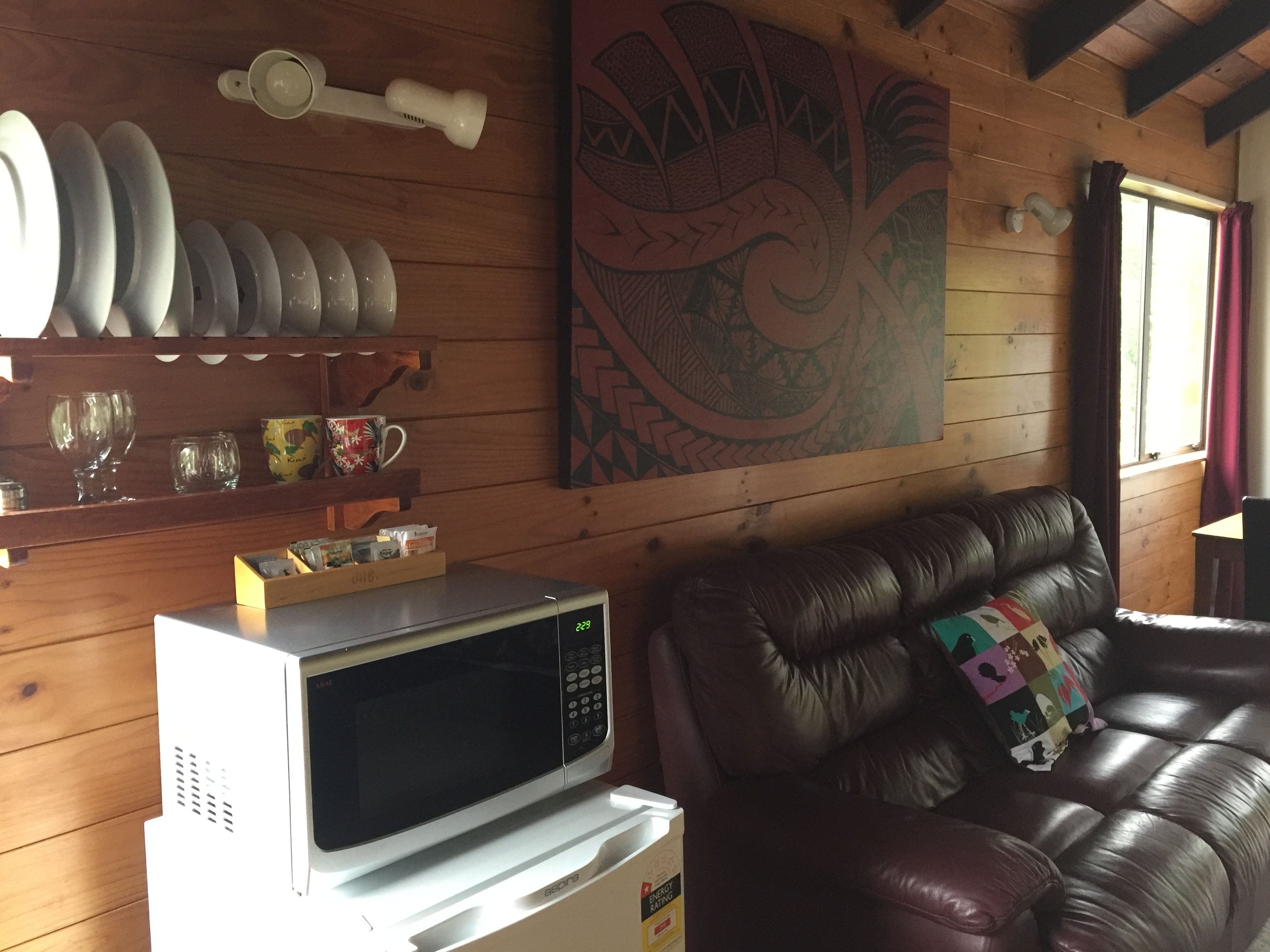 A cozy living room with a microwave on a counter, a brown leather couch, and a shelf with plates and cups.