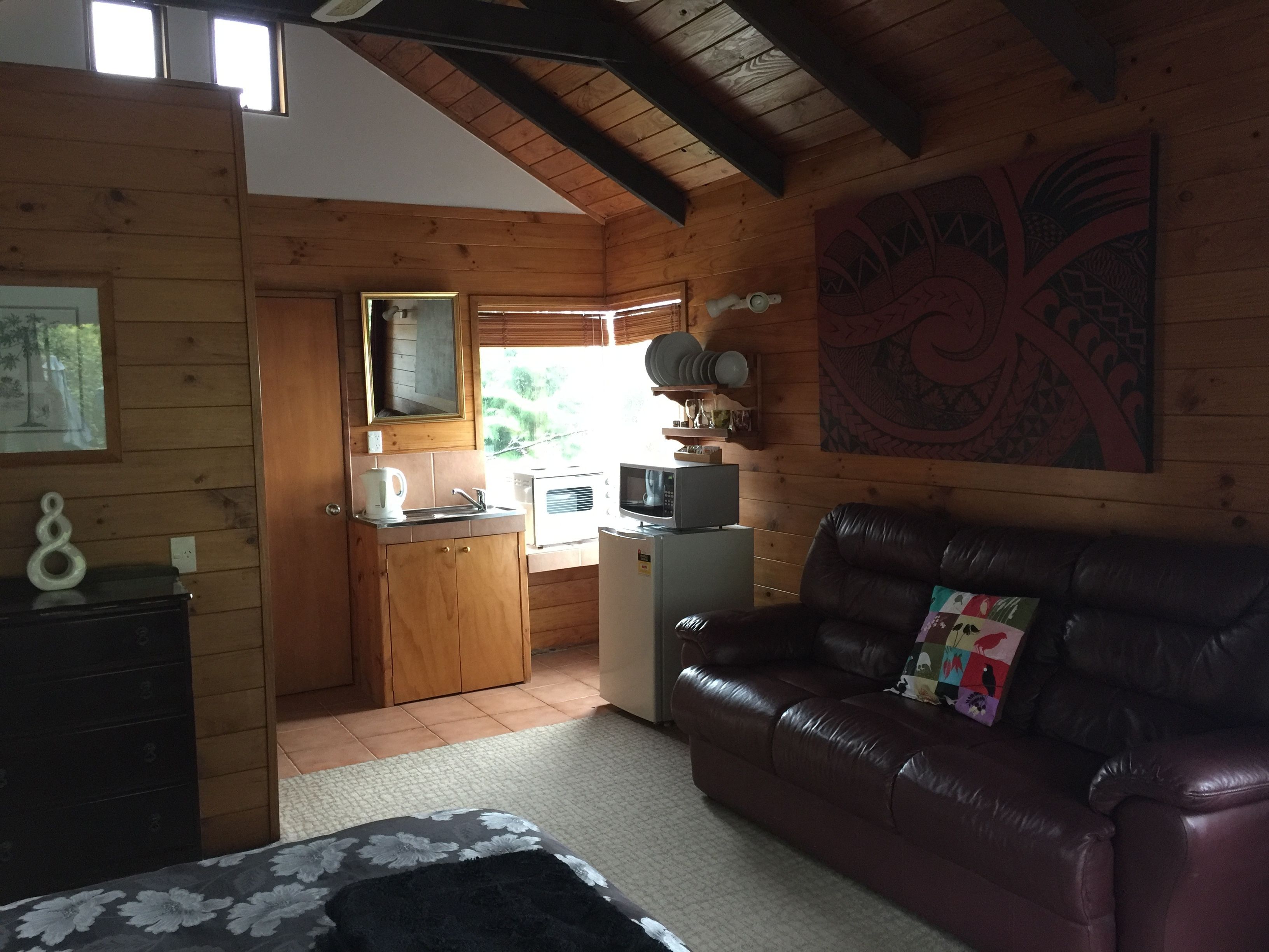A cozy cabin-style room with a wooden interior, a kitchenette, a leather sofa, and a large window providing natural light.