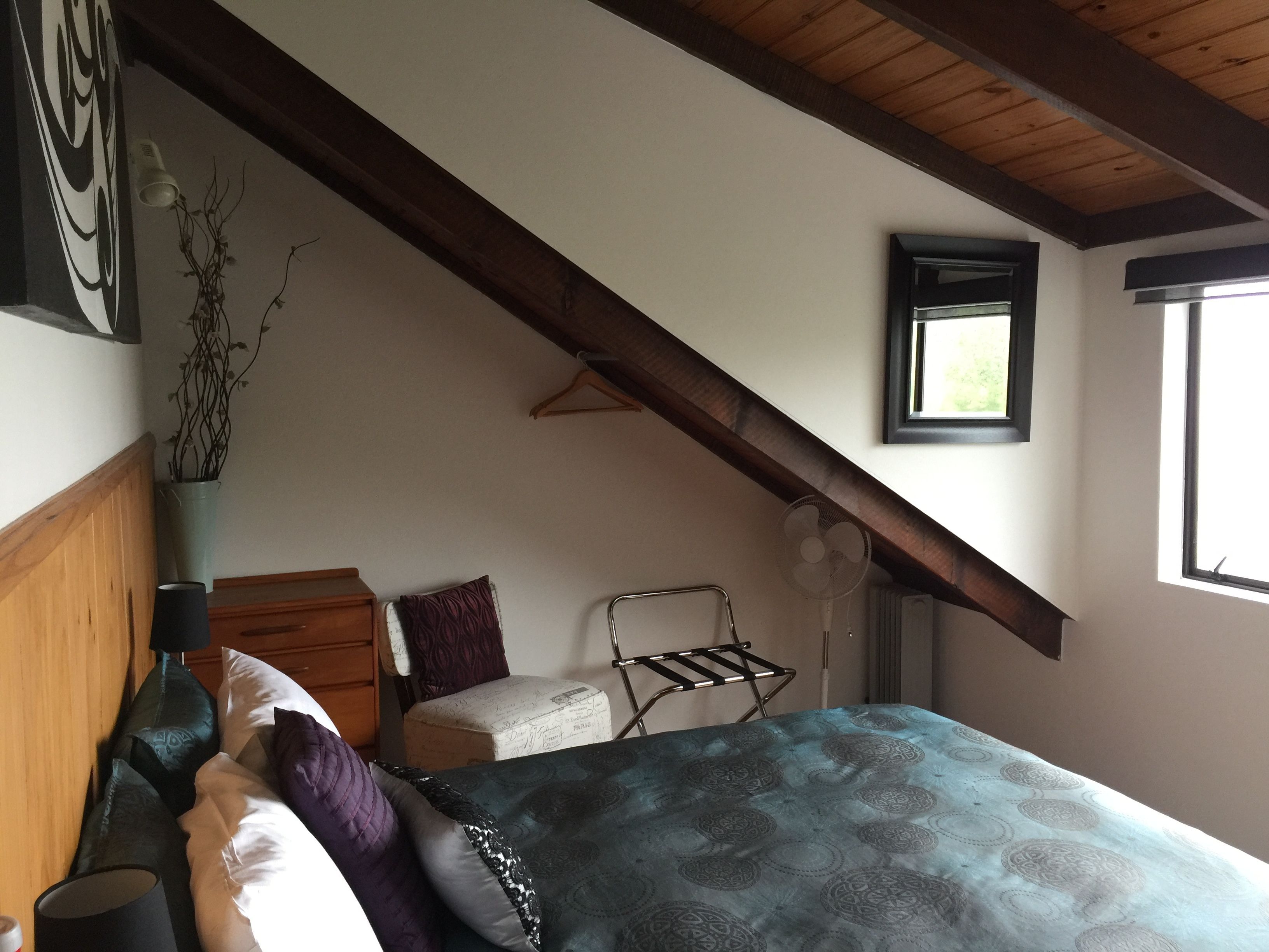 A bedroom with a sloped ceiling, a bed with patterned bedding, a bedside table, a hanging light fixture, and a window with a view of greenery.