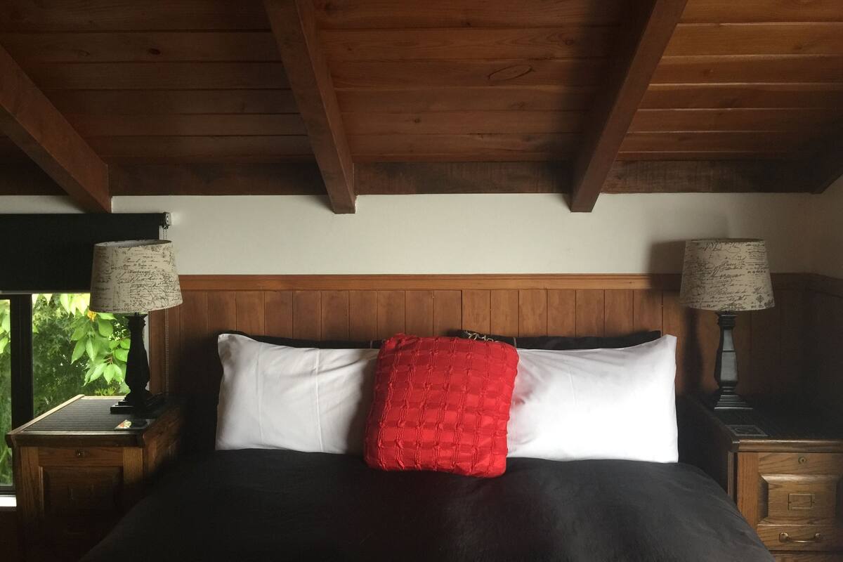 Un dormitorio con techo de madera, una cama con ropa de cama blanca y negra, un almohón rojo y dos mesitas de noche con lámparas.