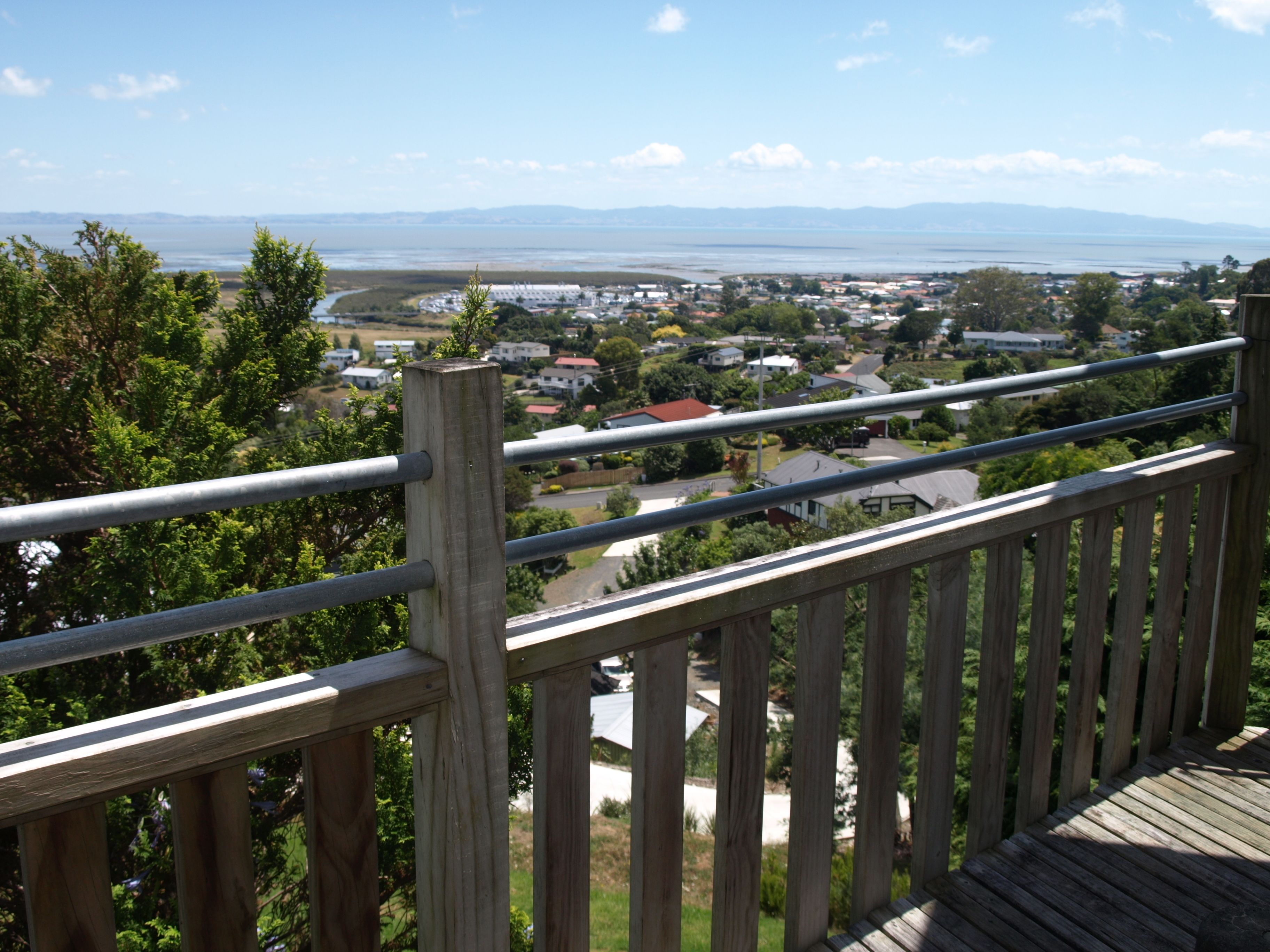 A wooden deck with a metal railing overlooking a residential area with houses and a body of water in the distance.