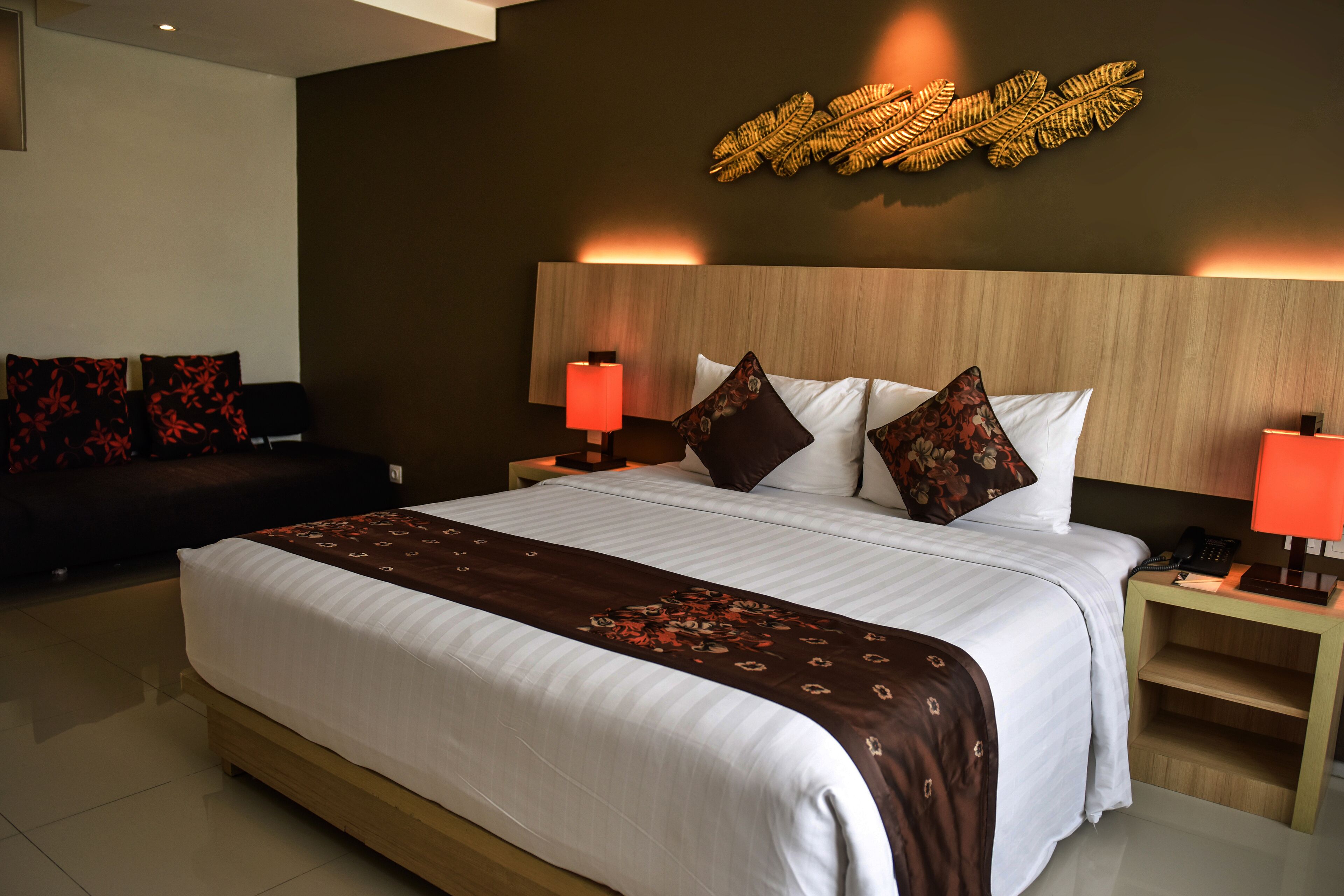 A hotel room with a large bed, two bedside tables with lamps, a dark sofa, and a decorative wall piece.