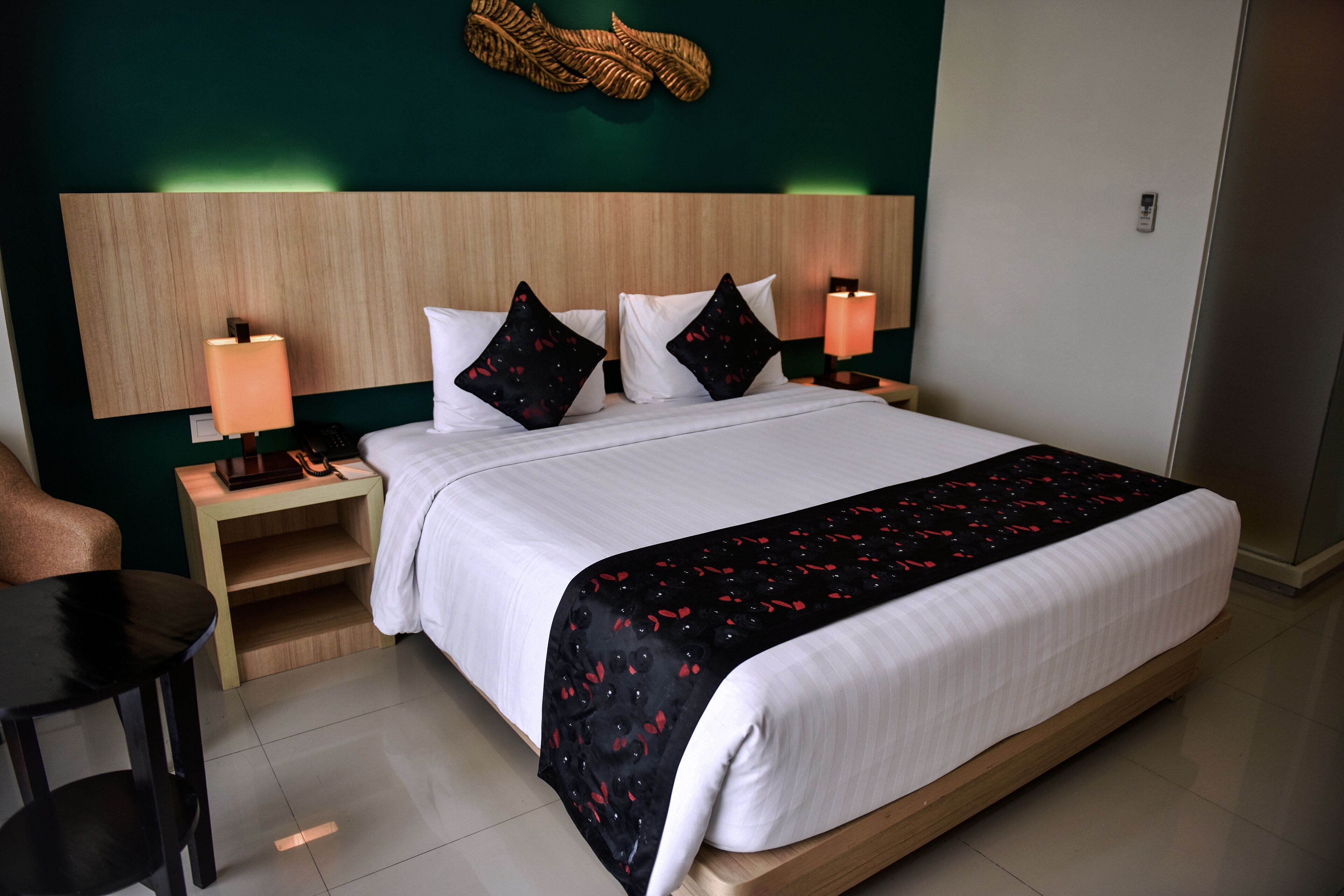 A neatly made bed with a black and red patterned bedspread, two bedside lamps, a wooden nightstand, and a wall-mounted decorative leaf.