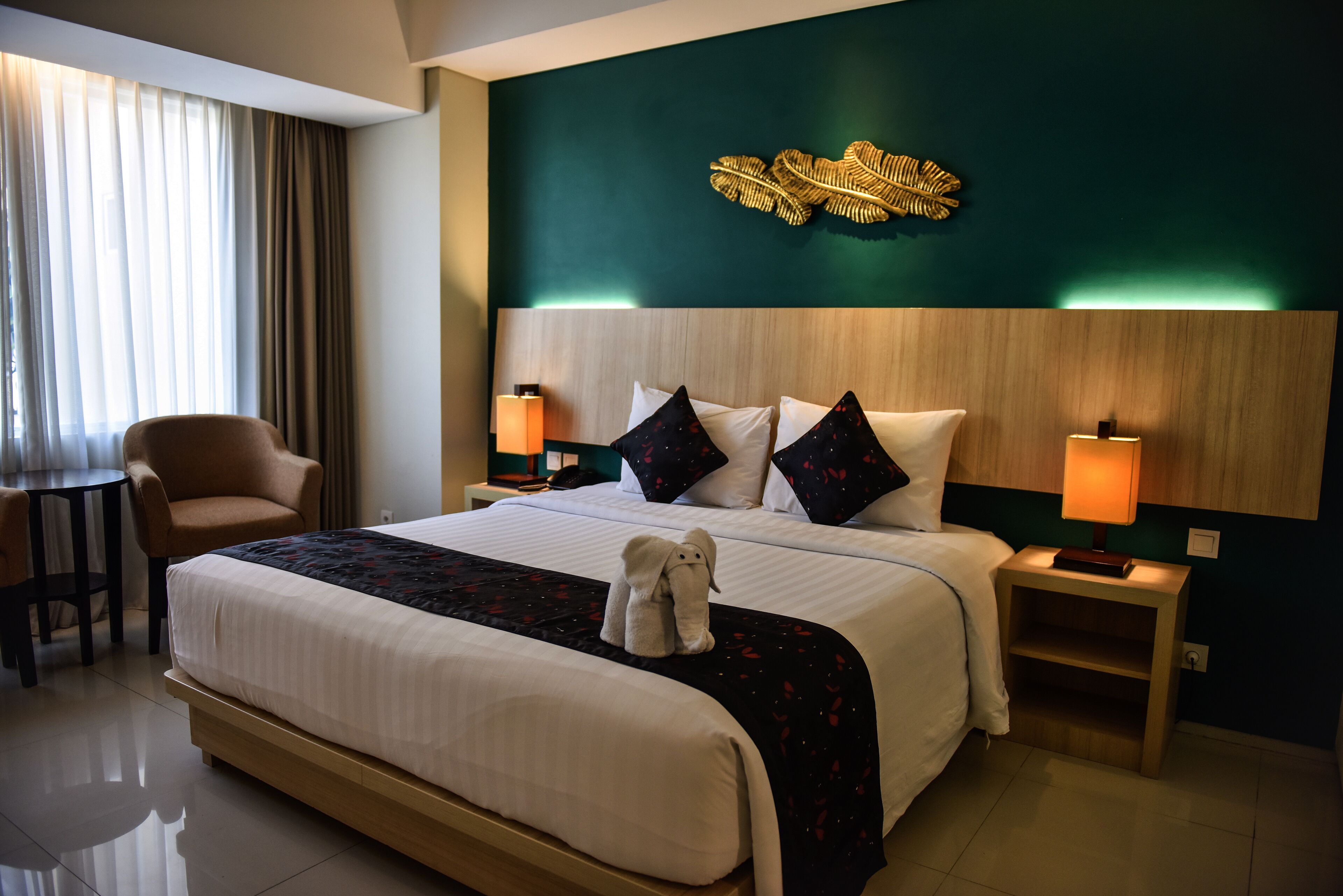 Ein Hotelzimmer mit einem großen Bett, zwei Nachttischen mit Lampen, einem Sessel, einem Schreibtisch und einer Wanddekoration mit Blättern.