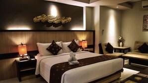 A modern hotel room with a large bed, two bedside tables with lamps, a sofa, and a decorative wall piece. - The Kana Kuta (Kuta)