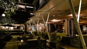 An outdoor seating area with tables and chairs, surrounded by potted plants, under a covered structure with a modern design. - The Kana Kuta (Kuta)