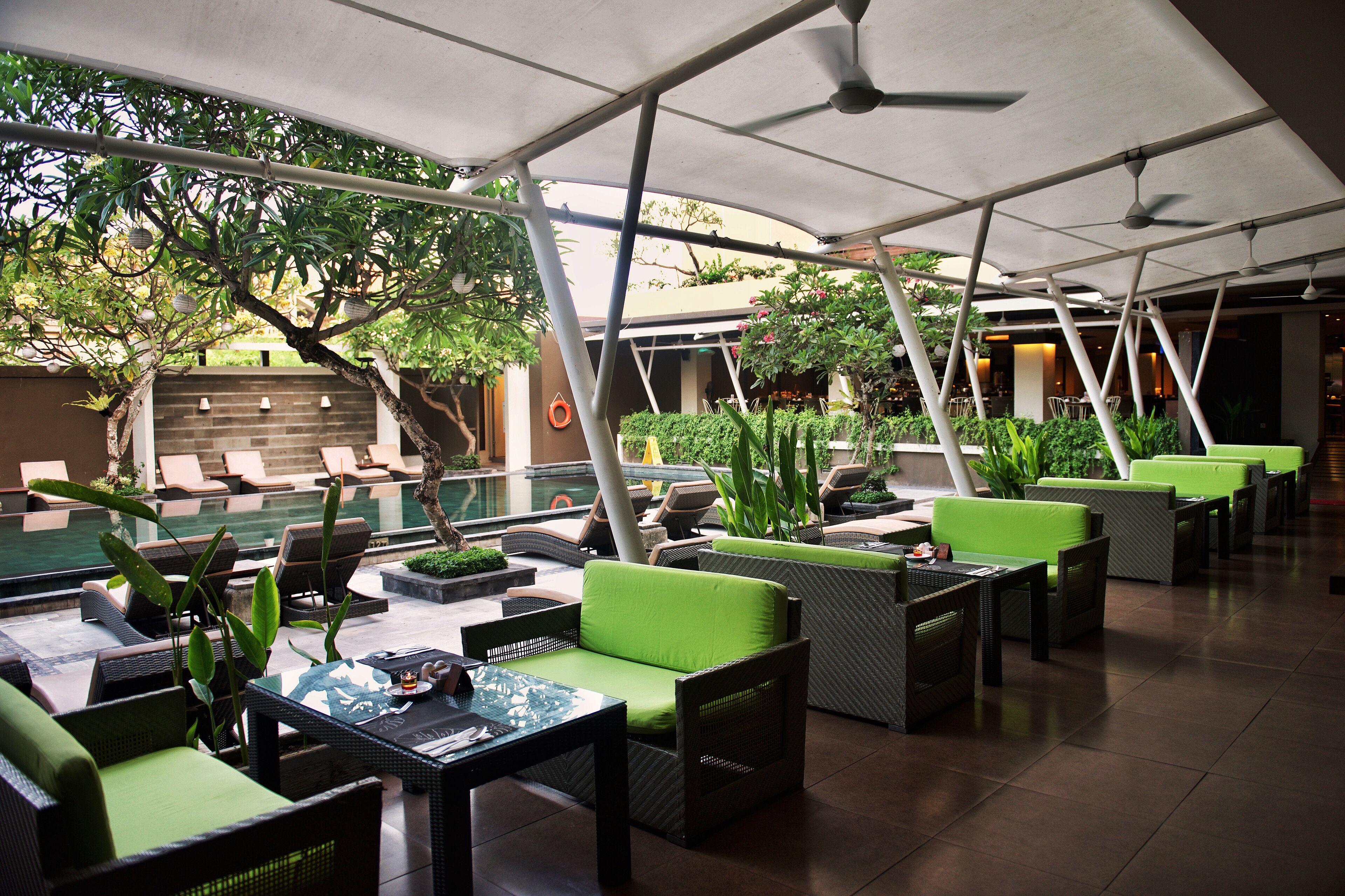 Een buitenzitgedeelte met groene, beklede stoelen en tafels, een zwembad op de achtergrond en een overdekte constructie met plafondventilatoren.