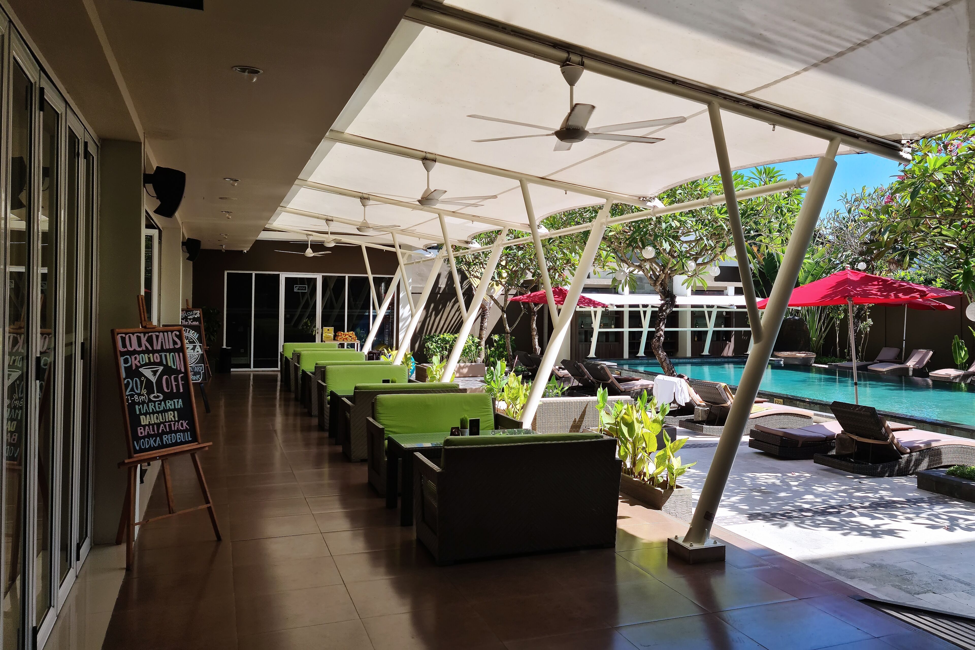A covered outdoor seating area with tables and chairs, a pool in the background, and a sign advertising cocktails promotion.
