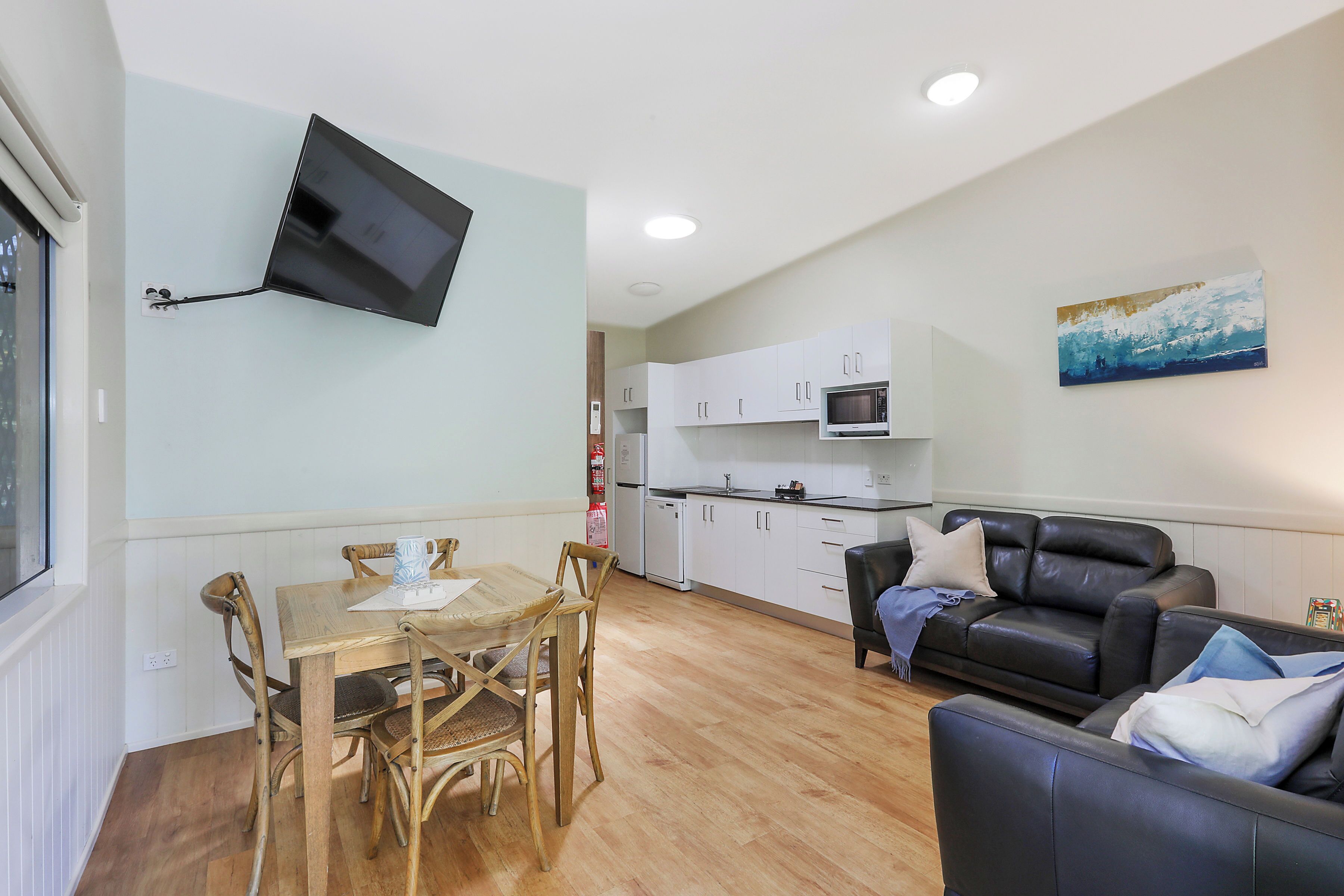 A compact living space with a dining area, a sofa, a flat-screen TV mounted on the wall, a kitchen with white cabinets, and a painting on the wall.