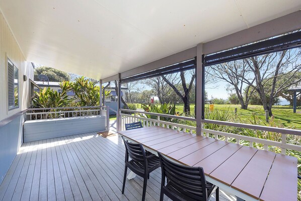 A covered outdoor area with a wooden table and chairs, a white railing, and a view of a garden and trees.