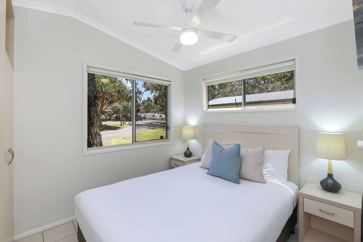A bedroom with a bed, two bedside tables, a ceiling fan, and two windows.