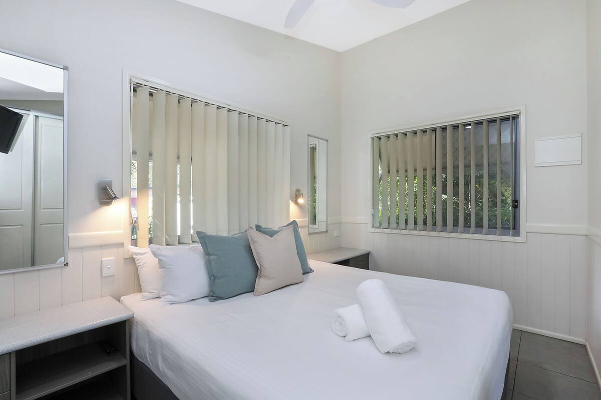 A hotel room with a bed, bedside tables, a mirror, and windows with blinds.