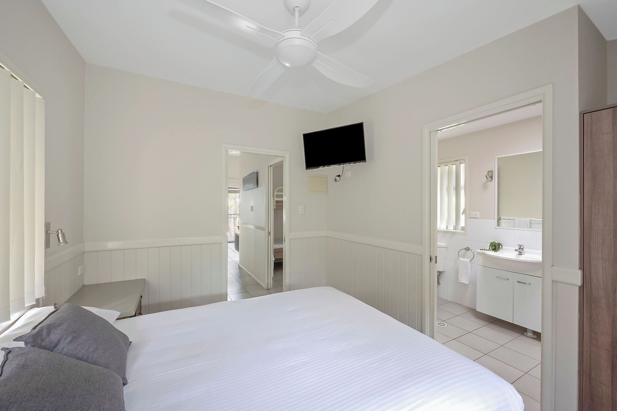 Une chambre à coucher avec un lit, un ventilateur de plafond, un téléviseur fixé au mur et une salle de bain visible à travers une porte ouverte.
