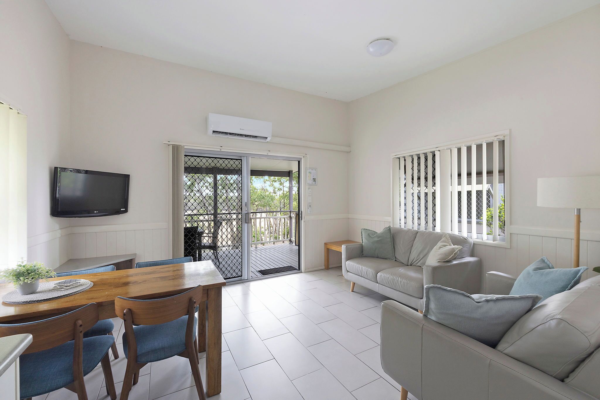 A spacious living area with a wooden dining table, blue chairs, a grey sofa, and a TV mounted on the wall.
