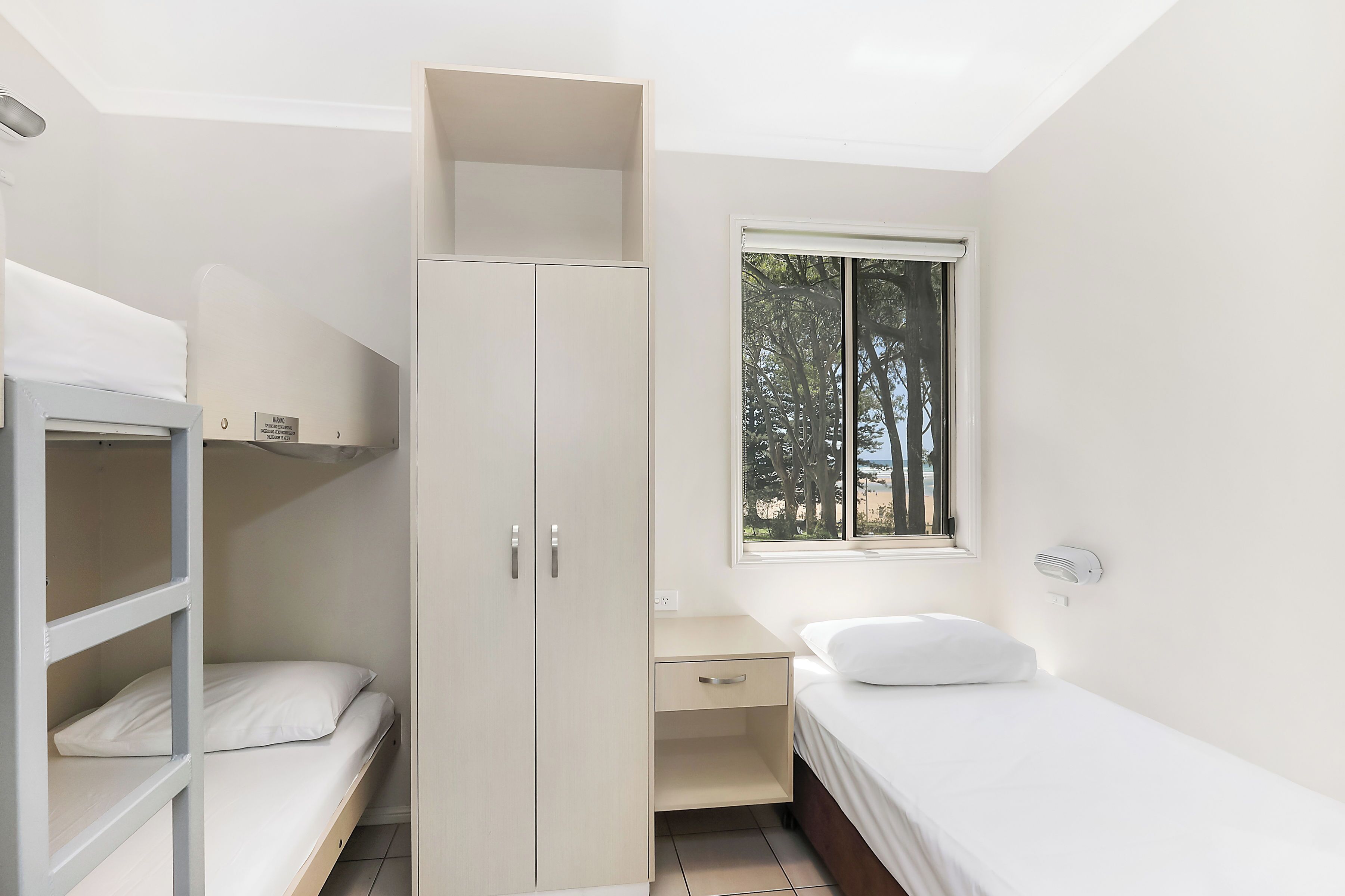 Une chambre compacte comprenant un lit superposé, un lit simple, une armoire, un bureau et une fenêtre donnant sur des arbres.