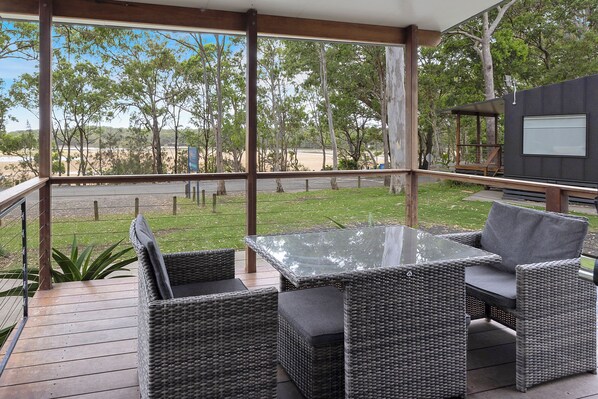 Un patio al aire libre techado con mobiliario de mimbre y una mesa de vidrio, con vistas a un área verde y árboles.