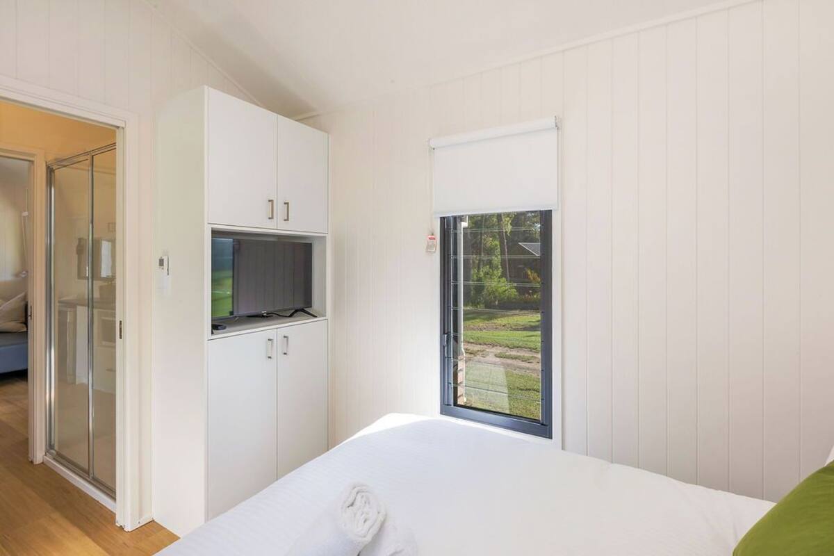 A bedroom with a bed, a TV on a cabinet, and a window with a view of greenery.