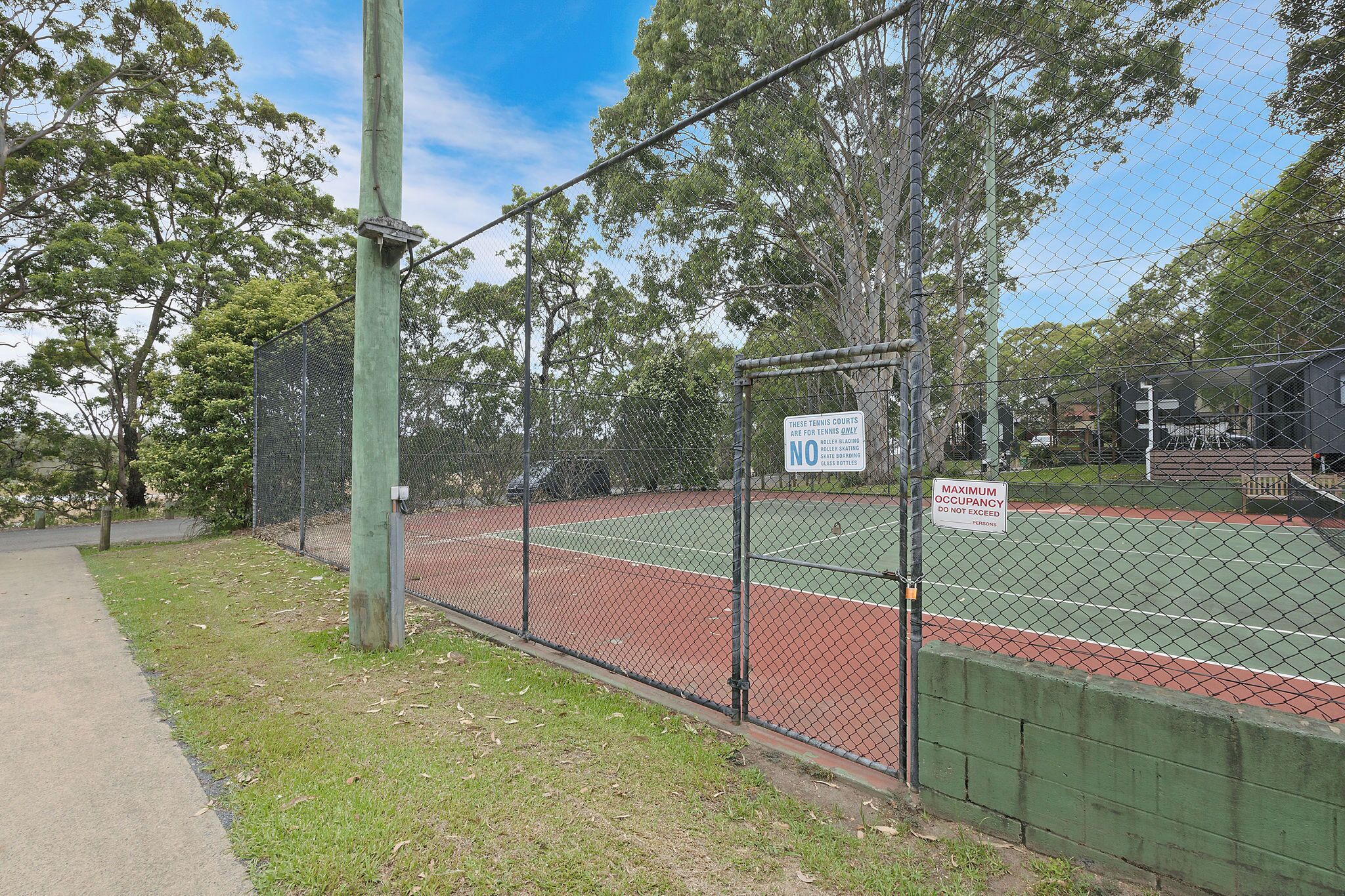 A fenced tennis court with a green fence, a sign indicating maximum occupancy, and trees in the background.