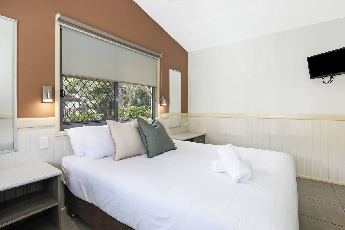 A hotel room with a large bed, a TV mounted on the wall, and a window with a view of greenery.