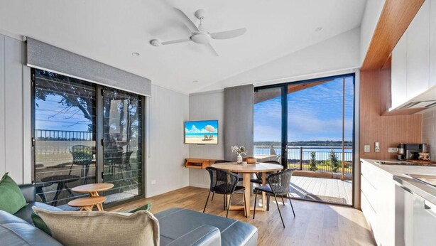 A modern living room with a sofa, a dining table, and chairs. There is a large window with a view of the outdoors. - Reflections Urunga - Holiday Park (Urunga)