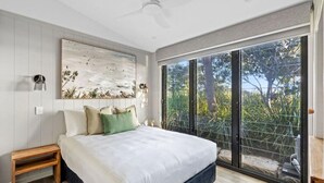 A bedroom with a large bed, a ceiling fan, a wall-mounted artwork, and a sliding glass door leading to a balcony with greenery. - Reflections Urunga - Holiday Park (Urunga)