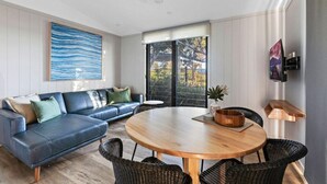 A modern living room with a blue sofa, a wooden dining table, wicker chairs, and a large window with a view. - Reflections Urunga - Holiday Park (Urunga)