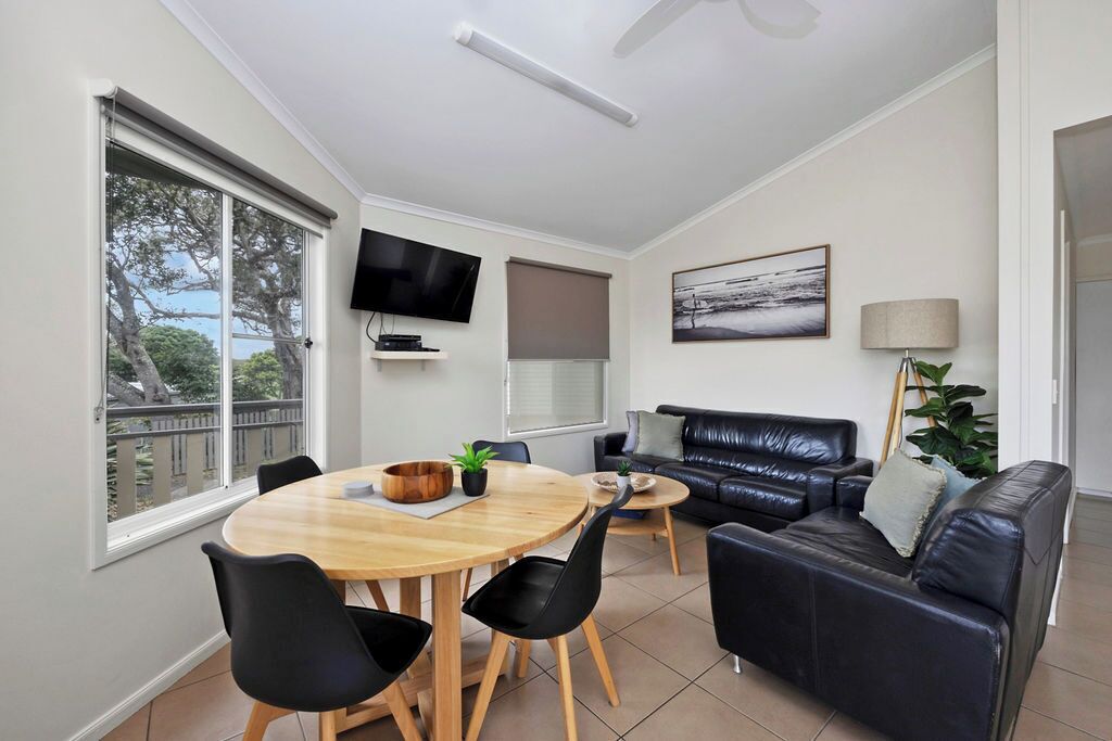 A modern living room with a round wooden dining table, black chairs, a black leather sofa, a TV mounted on the wall, and a large window with a view of trees.