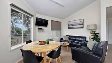 A modern living room with a round wooden dining table, black chairs, a black leather sofa, a TV mounted on the wall, and a large window with a view of trees.