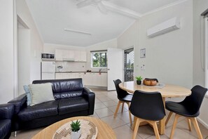 A modern living room with a black leather sofa, a wooden dining table with black chairs, and a kitchen in the background.