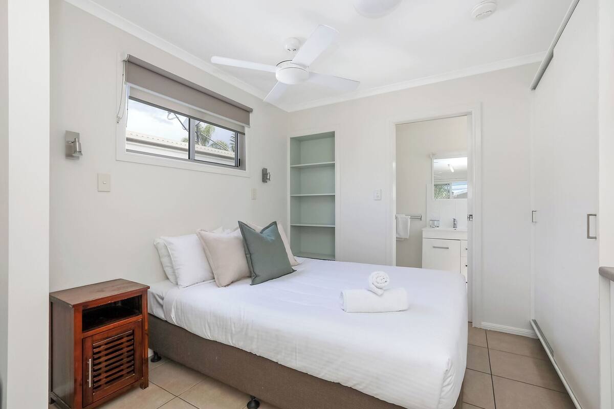 A bedroom with a bed, a ceiling fan, a window with blinds, a bedside table, and a built-in shelf.