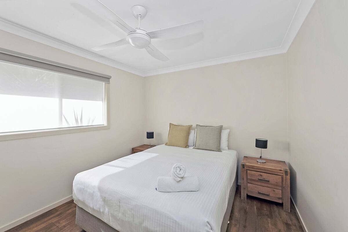 A bedroom with a bed, a ceiling fan, a nightstand with a lamp, and a window with blinds.