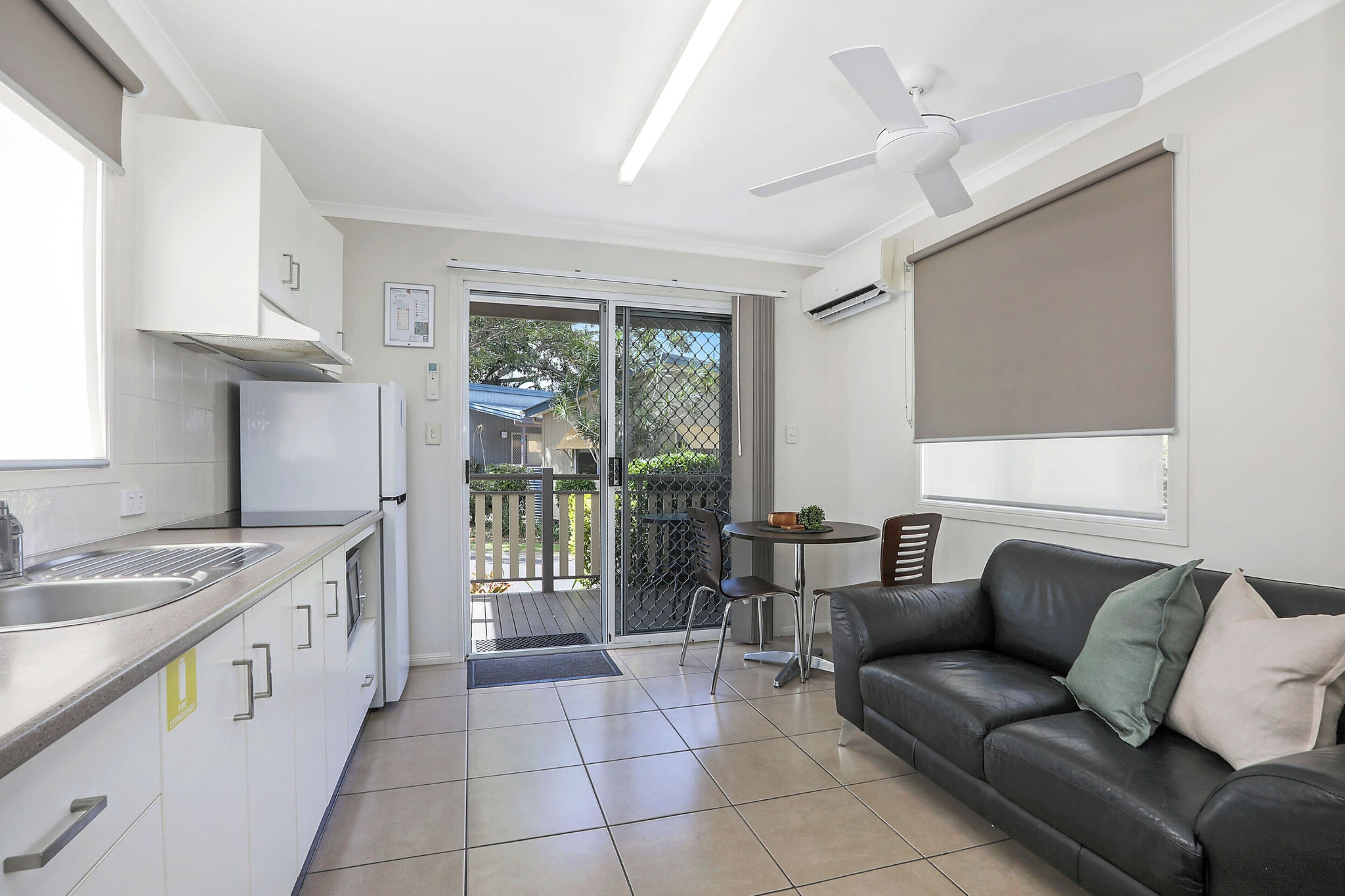A modern kitchen with a refrigerator, sink, and cabinets. There is a dining area with a table and chairs, and a black leather sofa. A ceiling fan and air conditioning unit are visible.