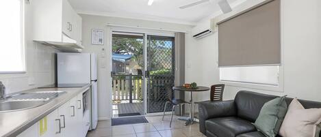 Een moderne keuken met een koelkast, spoelbak en kasten. Er is een eethoek met een tafel en stoelen, en een zwarte leren bank. Een plafondventilator en airconditioning zijn zichtbaar.