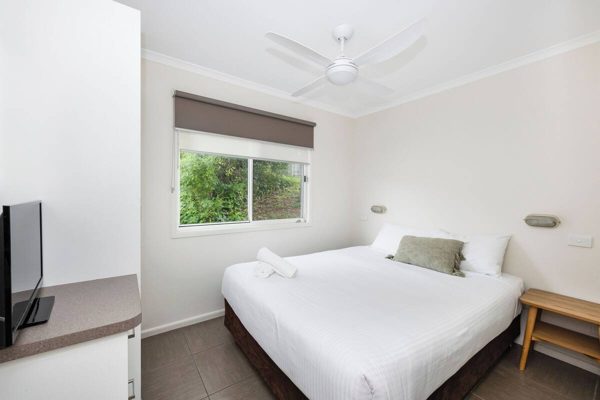 A bedroom with a bed, a window with a view of greenery, a ceiling fan, and a bedside table.
