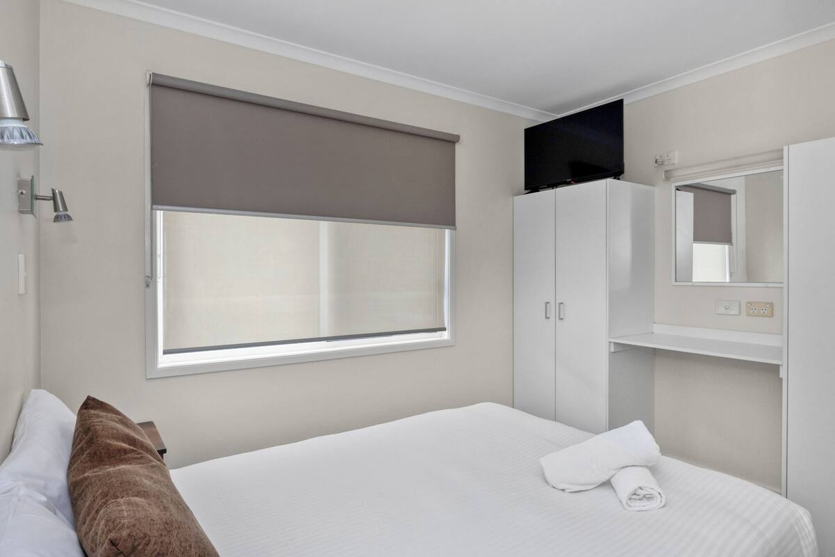 A hotel room with a bed, a window with a blind, a TV mounted above the bed, and a mirror above the door.