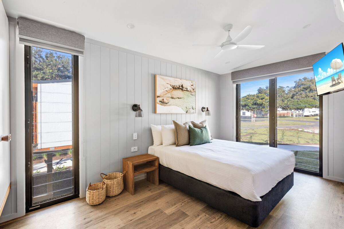 A bedroom with a large bed, a ceiling fan, a TV, and a view of the outdoors.