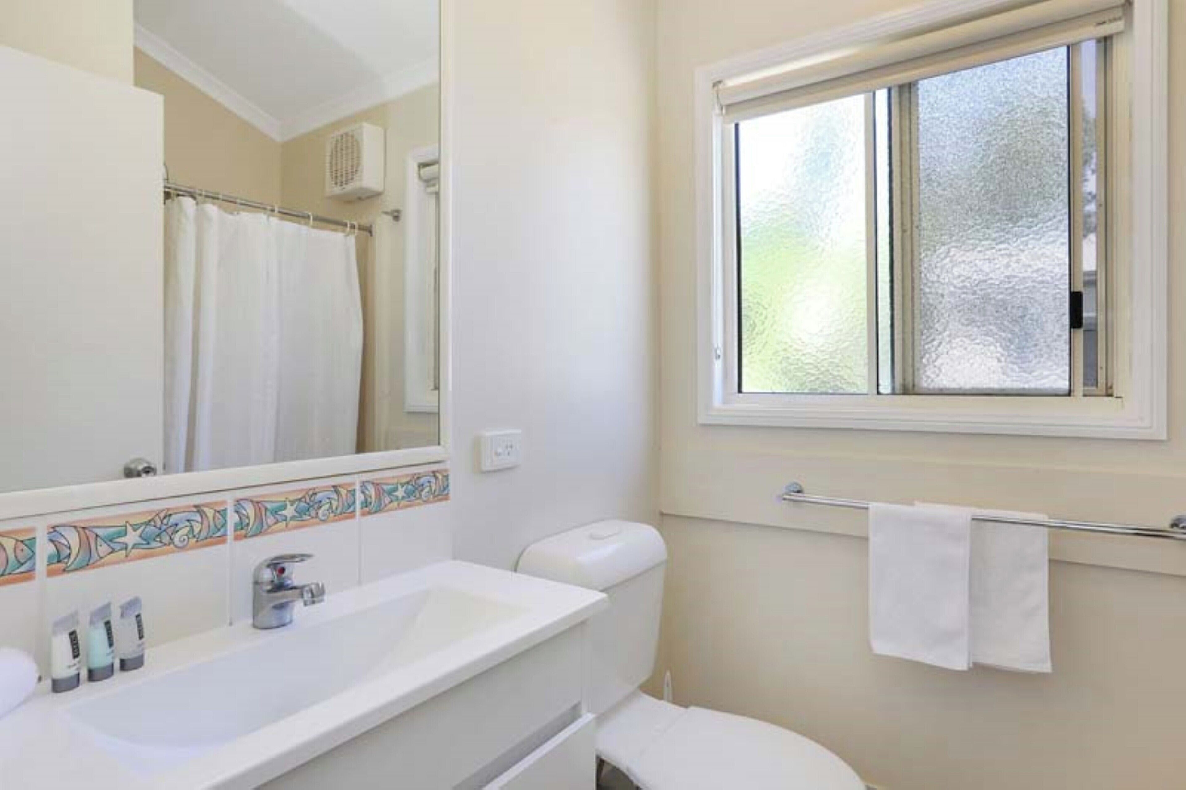 A bathroom with a white sink, mirror, and toilet. There is a window with a frosted glass pane and a towel hanging on a rack.