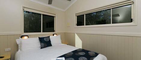 A hotel room with a bed, two windows, a ceiling fan, and a bedside table with a lamp.
