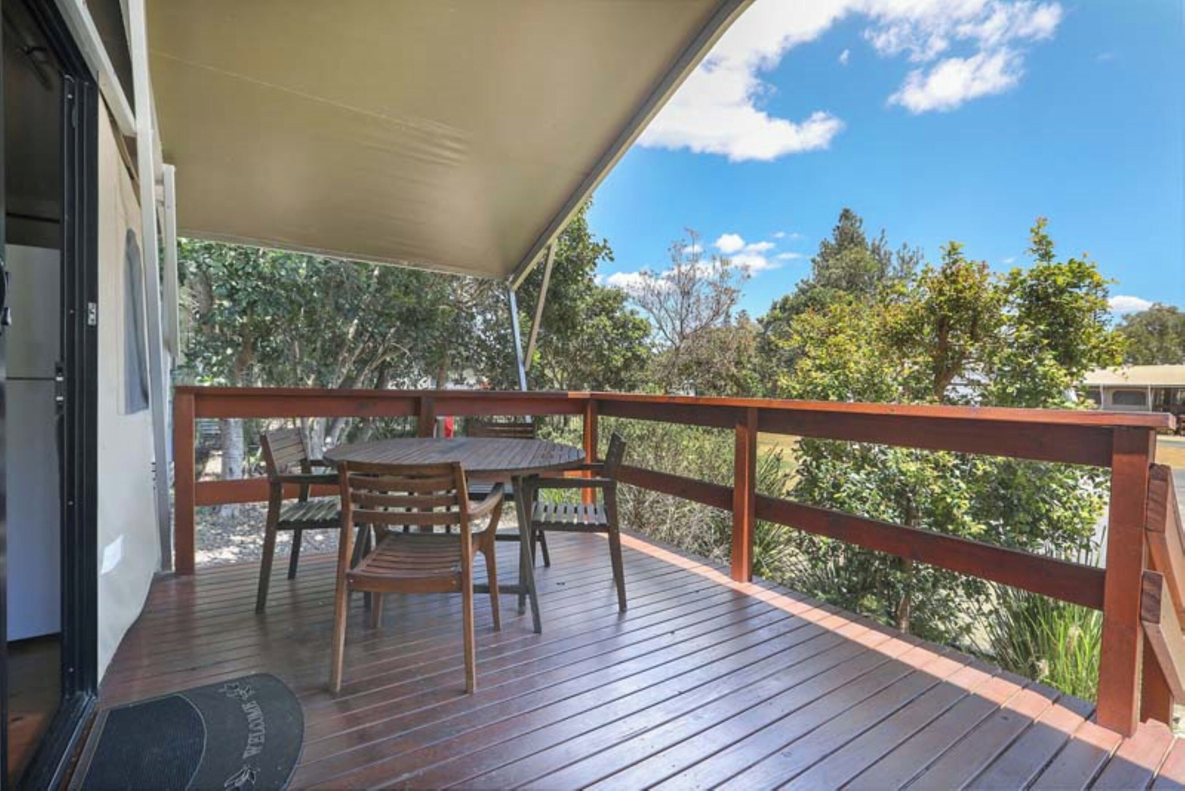 Un patio in legno con tavolo e sedie, una zona riparata e vista su alberi e cielo.