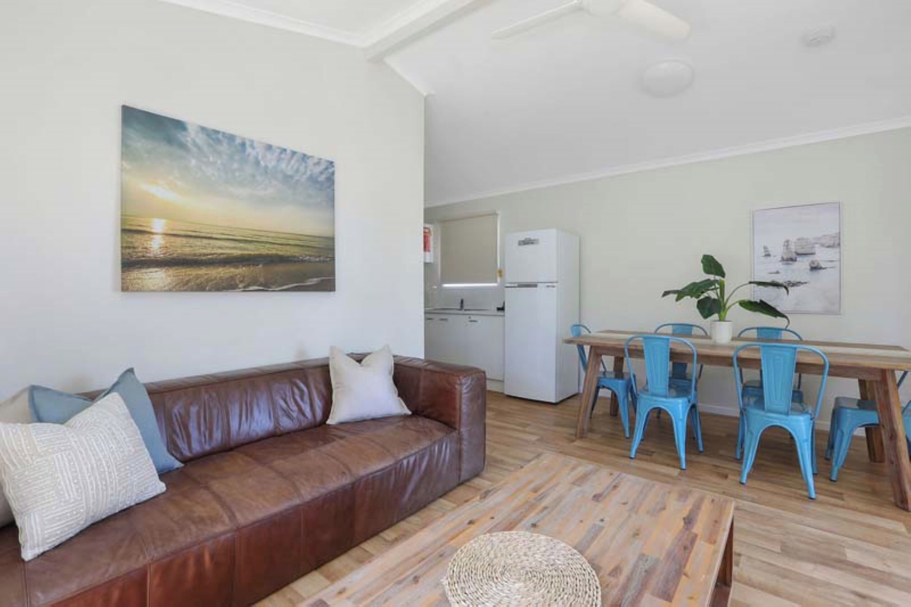 Una sala de estar con un sofá de cuero marrón, una mesa de comedor con sillas azules y una zona de cocina con un refrigerador blanco.