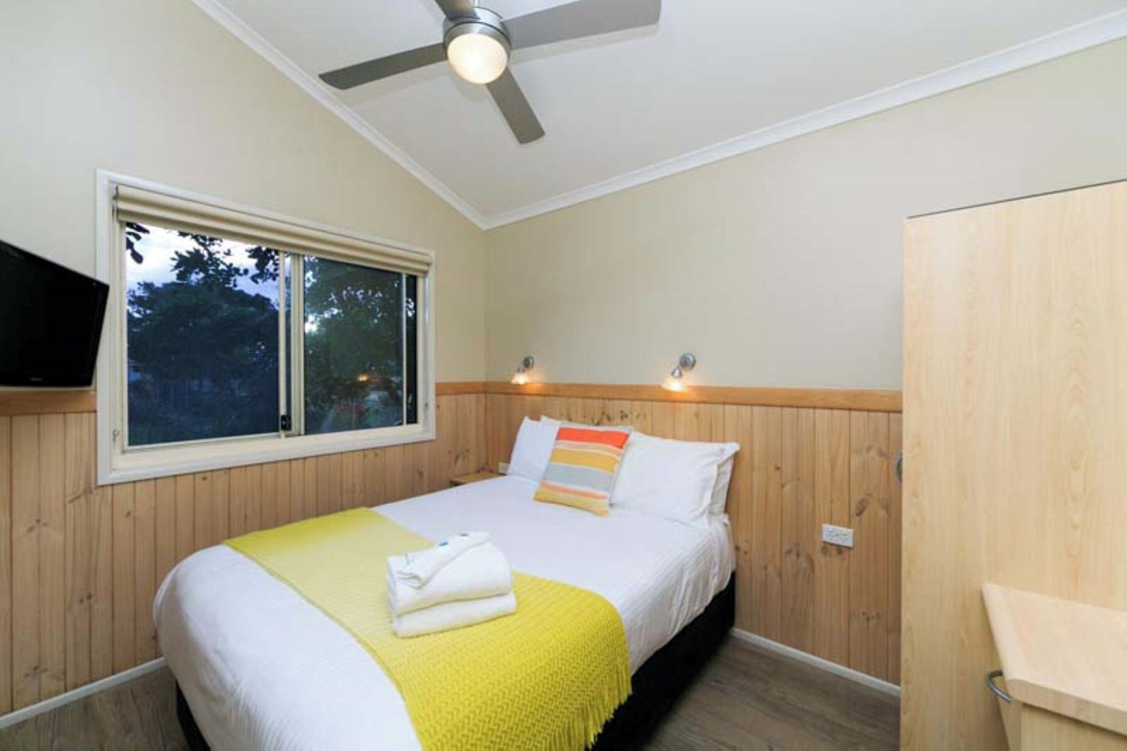 Un dormitorio con cama, ventilador de techo, ventana y televisor montado en la pared.
