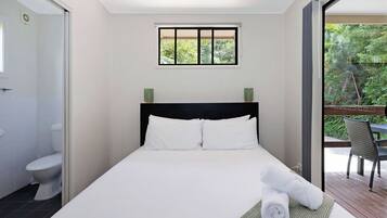 A hotel room with a bed, a window with a view of greenery, a bathroom with a toilet, and a balcony with a chair.