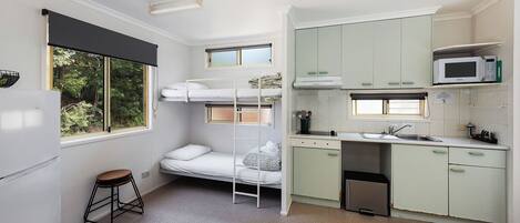 A compact room with bunk beds, a kitchenette, a small stool, and a window with a view of greenery.