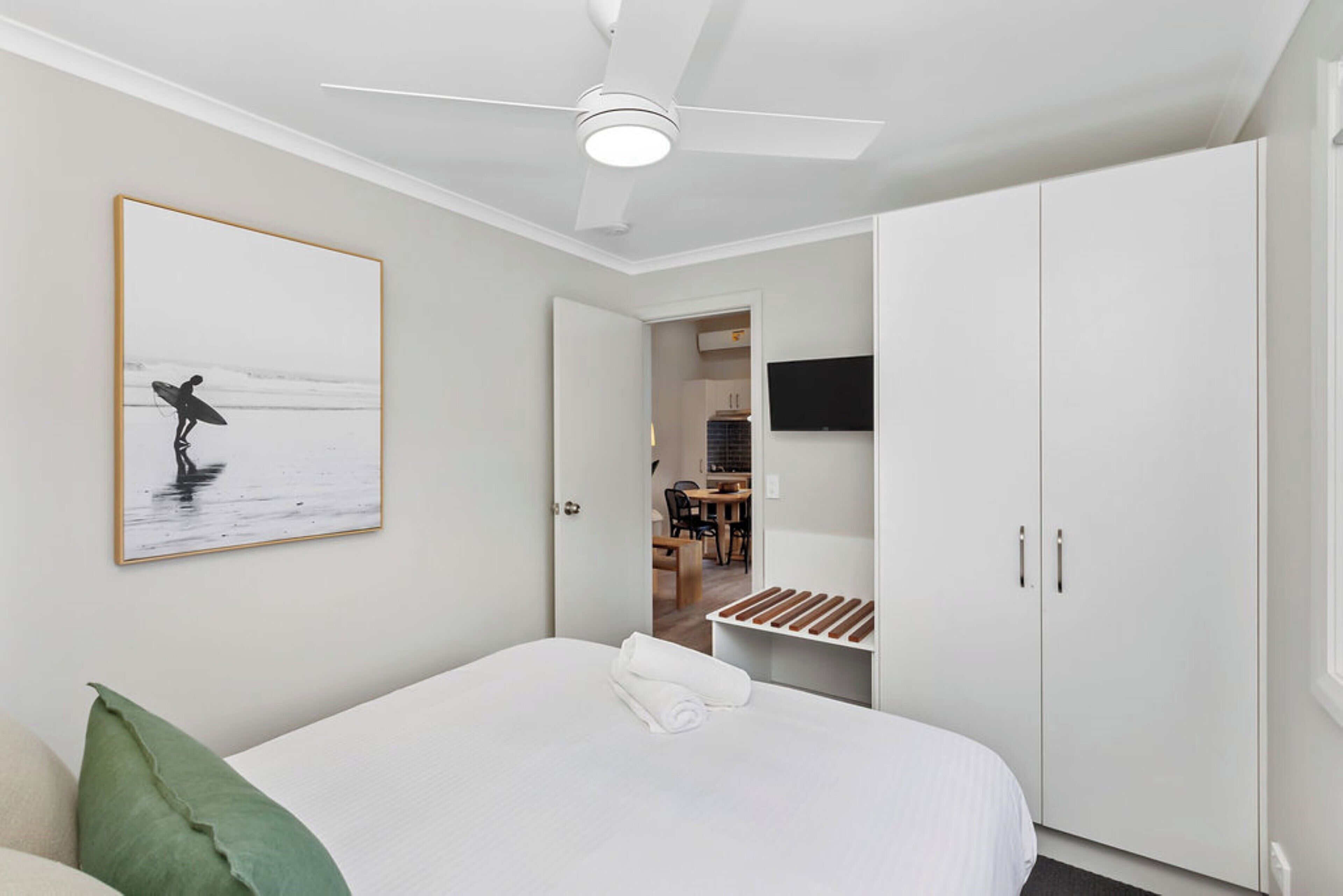 A bedroom with a bed, a ceiling fan, a framed picture, a wardrobe, and a view into the dining area.