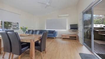 Una sala de estar moderna con una mesa de comedor de madera, sillas negras, un sofá azul, un televisor y un ventanal corredizo que da a un espacio exterior.